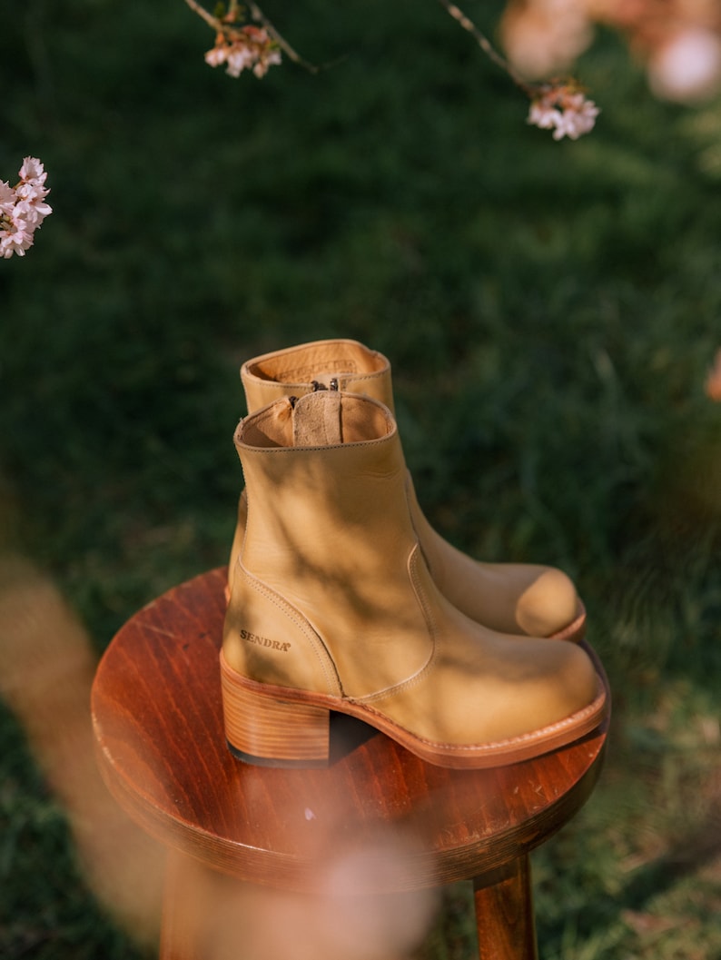 May include: Tan leather ankle boots with a chunky heel and a side zip. The boots are sitting on a wooden stool. The brand name "SENDRA" is visible on the side of the boot.