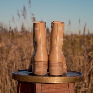 May include: A pair of tan leather ankle boots with white daisy embroidery. The boots are sitting on a wooden stool with a brass top.