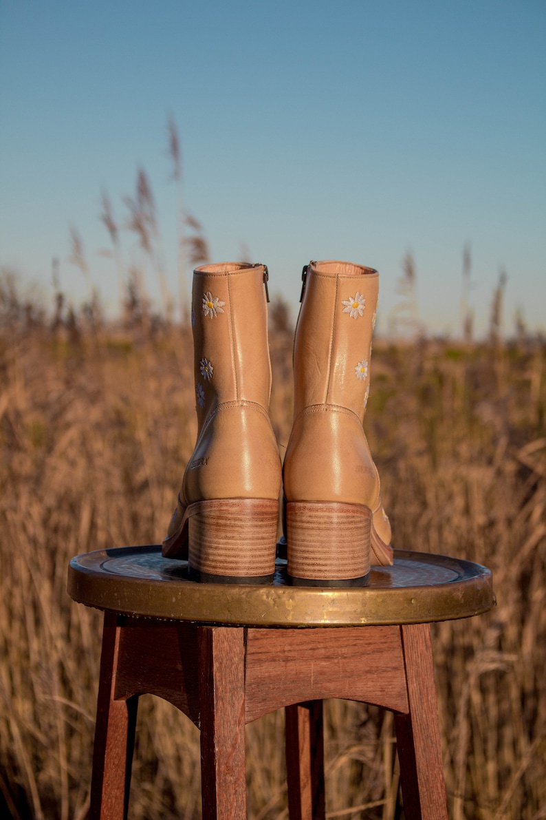 May include: A pair of tan leather ankle boots with white daisy embroidery. The boots are sitting on a wooden stool with a brass top.