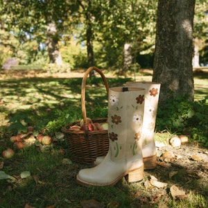 May include: A pair of beige knee-high boots with floral embroidery. The boots are sitting on a bed of fallen leaves and apples in a grassy area.