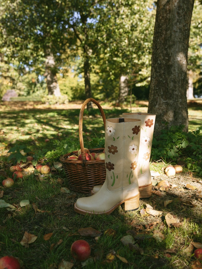 May include: A pair of beige knee-high boots with floral embroidery. The boots are sitting on a bed of fallen leaves and apples in a grassy area.