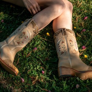 Może przedstawiać: Para beżowych sk&oacute;rzanych kowbojskich but&oacute;w z kwiatowymi haftami i wzorem sk&oacute;ry węża na czubku. Buty mają gruby obcas i są noszone przez osobę siedzącą na trawiastym polu.
