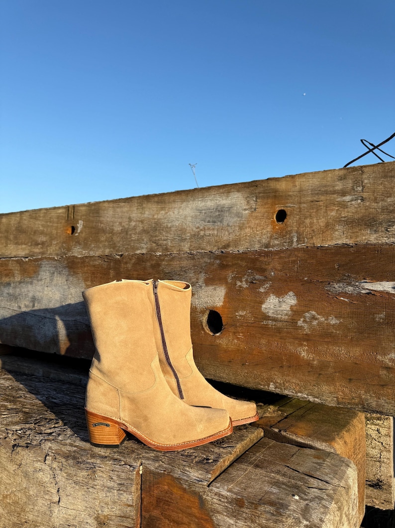 Puede incluir: Un par de botines de ante color beige con cierre de cremallera. Los botines tienen punta cuadrada, tac&oacute;n de madera apilada y suela roja. Se encuentran sobre vigas de madera desgastadas y un cielo azul claro.