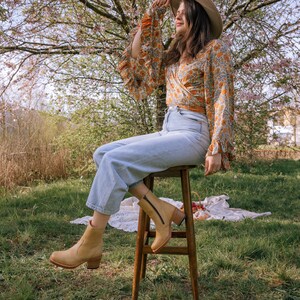 May include: A woman wearing a floral print blouse, blue jeans, and tan ankle boots sits on a wooden stool in a grassy field. She is wearing a wide-brimmed hat and has her hand on the brim.