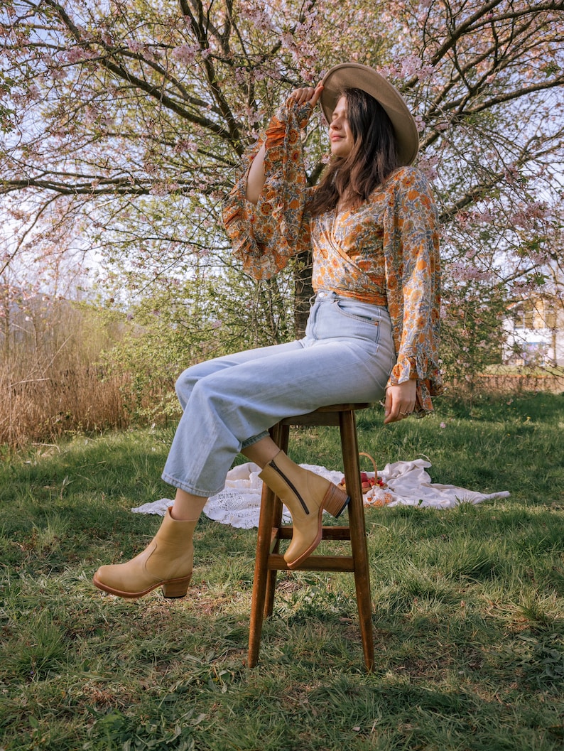May include: A woman wearing a floral print blouse, blue jeans, and tan ankle boots sits on a wooden stool in a grassy field. She is wearing a wide-brimmed hat and has her hand on the brim.