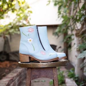 Może przedstawiać: Para jasnoniebieskich kozaków za kostkę z zamkiem błyskawicznym z boku. Kozaki są ozdobione haftowanymi kwiatami w kolorze różowym, białym i zielonym.