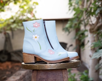 May include: A pair of light blue ankle boots with a zipper on the side. The boots are decorated with embroidered flowers in pink, white, and green.