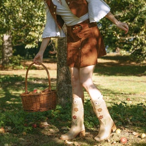 May include: A woman wearing a brown suede vest and skirt, white blouse, and floral print boots, carries a wicker basket filled with apples in a fall setting.