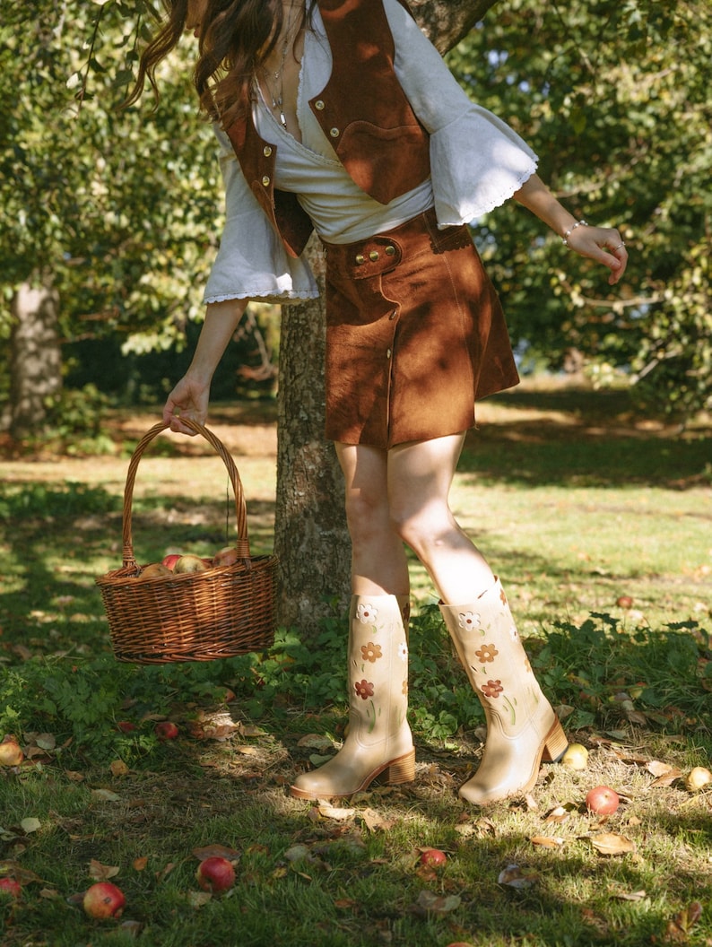 May include: A woman wearing a brown suede vest and skirt, white blouse, and floral print boots, carries a wicker basket filled with apples in a fall setting.