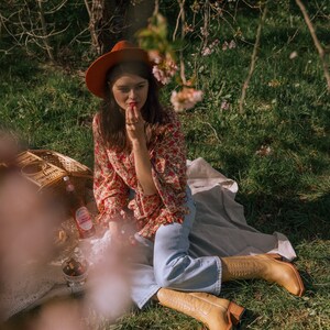 Puede incluir: Una persona con sombrero color &oacute;xido, blusa floral y vaqueros azul claro sentada en una manta de picnic. Lleva botas de vaquero color beige. Una cesta de picnic, bebidas y aperitivos son visibles en la manta, con hierba verde y &aacute;rboles al fondo.