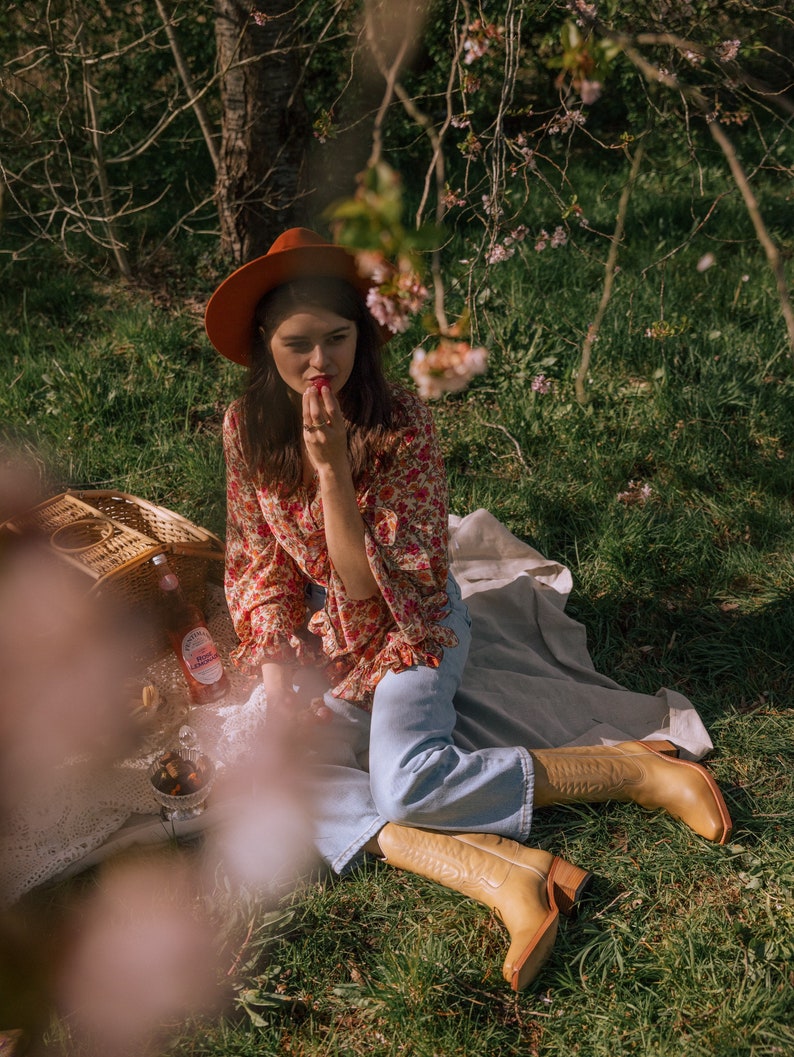 Puede incluir: Una persona con sombrero color &oacute;xido, blusa floral y vaqueros azul claro sentada en una manta de picnic. Lleva botas de vaquero color beige. Una cesta de picnic, bebidas y aperitivos son visibles en la manta, con hierba verde y &aacute;rboles al fondo.