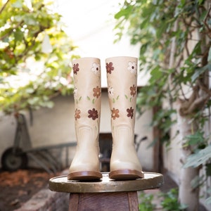 May include: A pair of beige leather boots with floral embroidery. The boots are sitting on a wooden stool.