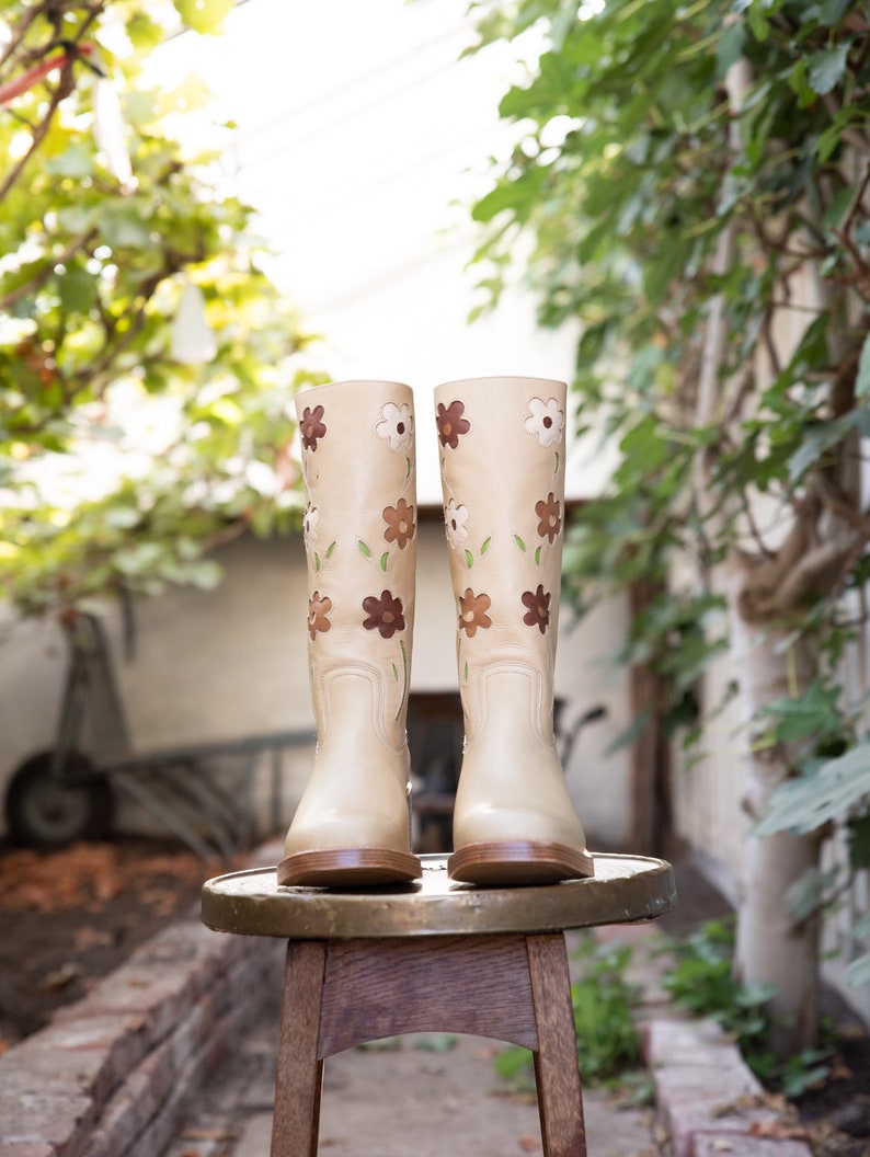 May include: A pair of beige leather boots with floral embroidery. The boots are sitting on a wooden stool.