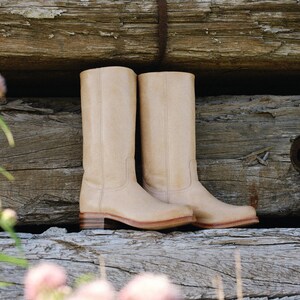 May include: A pair of beige leather cowboy boots with a classic design. The boots have a tall shaft, a low heel, and a rounded toe. They are set against a rustic wooden background.