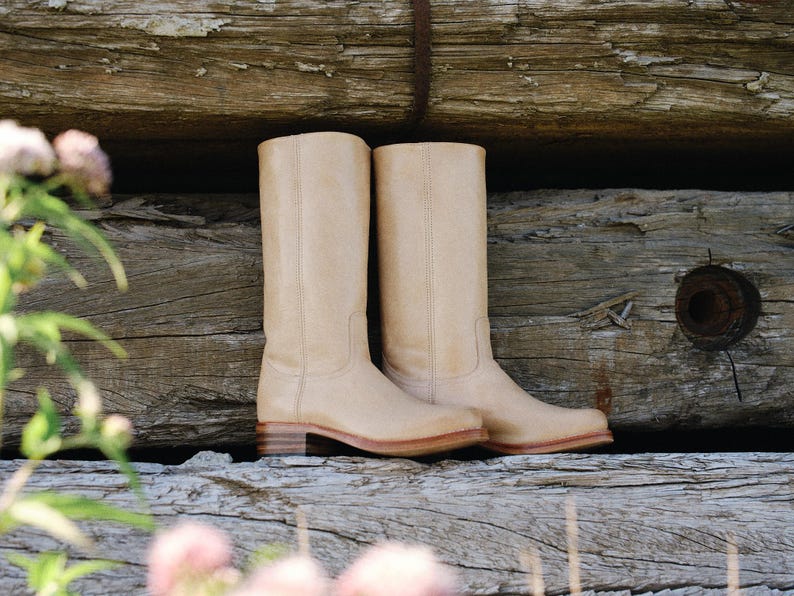 May include: A pair of beige leather cowboy boots with a classic design. The boots have a tall shaft, a low heel, and a rounded toe. They are set against a rustic wooden background.
