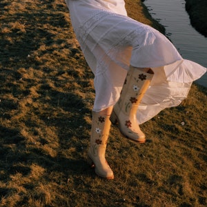 May include: A woman wearing a white lace dress and floral embroidered cowboy boots. The boots are a light brown color with white and brown flowers.