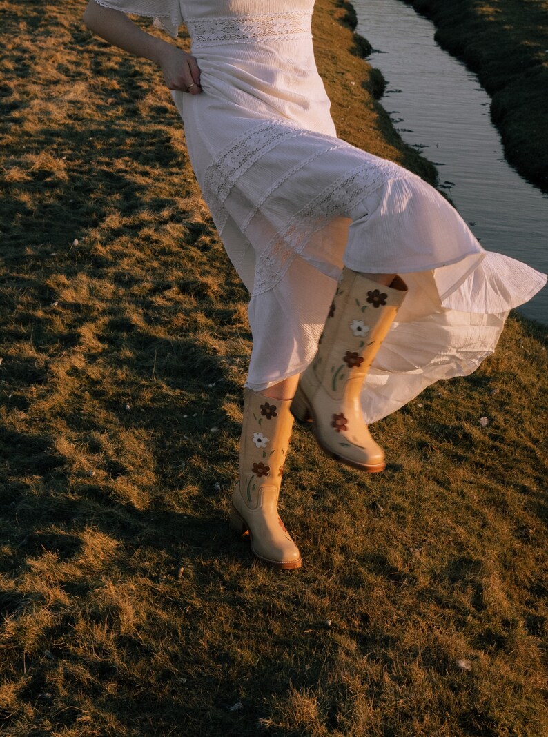 May include: A woman wearing a white lace dress and floral embroidered cowboy boots. The boots are a light brown color with white and brown flowers.