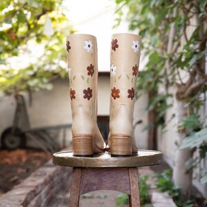 May include: A pair of beige leather knee-high boots with brown, white, and green floral embroidery. The boots are sitting on a wooden stool.