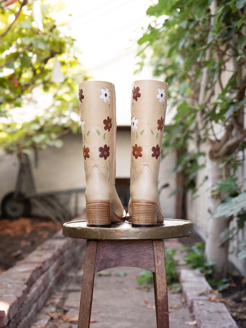 May include: A pair of beige leather knee-high boots with brown, white, and green floral embroidery. The boots are sitting on a wooden stool.