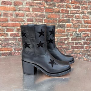 Może przedstawiać: Czarne skórzane botki z wycięciami w kształcie gwiazd. Botki mają obcas w kształcie klocka. Tło to ceglana ściana. Stylowe obuwie.