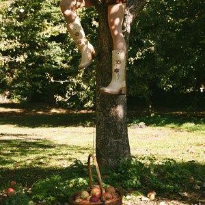 May include: A woman wearing a brown skirt and beige boots with floral embroidery climbs a tree. A wicker basket filled with red apples sits at the base of the tree.
