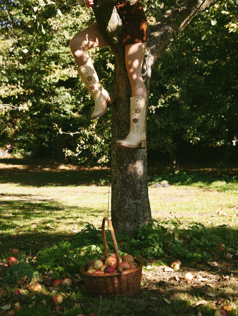 May include: A woman wearing a brown skirt and beige boots with floral embroidery climbs a tree. A wicker basket filled with red apples sits at the base of the tree.
