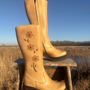 May include: Tan leather knee-high boots with a floral design cut out on the side. The boots have a chunky heel and a platform sole. The boots are sitting on a wooden stool.