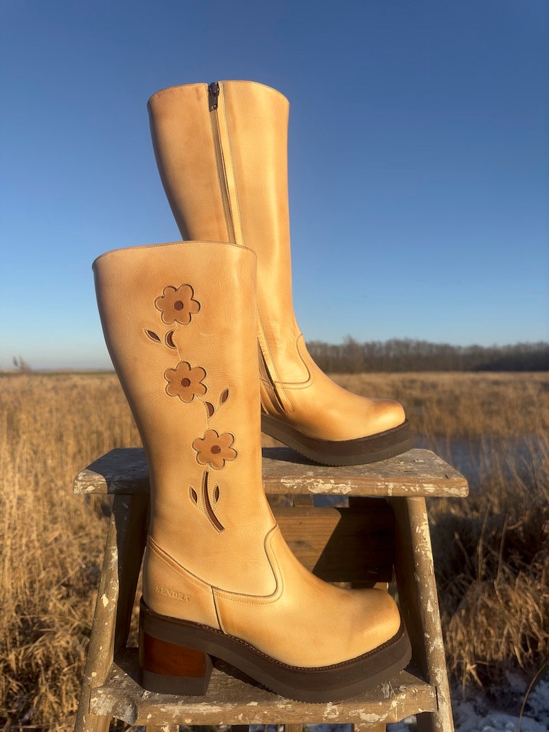 May include: Tan leather knee-high boots with a floral design cut out on the side. The boots have a chunky heel and a platform sole. The boots are sitting on a wooden stool.