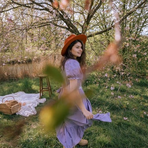 May include: A woman wearing a rust-colored hat and a lavender dress stands in a grassy area with a picnic basket and blanket. The scene is framed by a flowering tree, creating a soft, natural aesthetic. The dress has a ruffled hem and short sleeves.