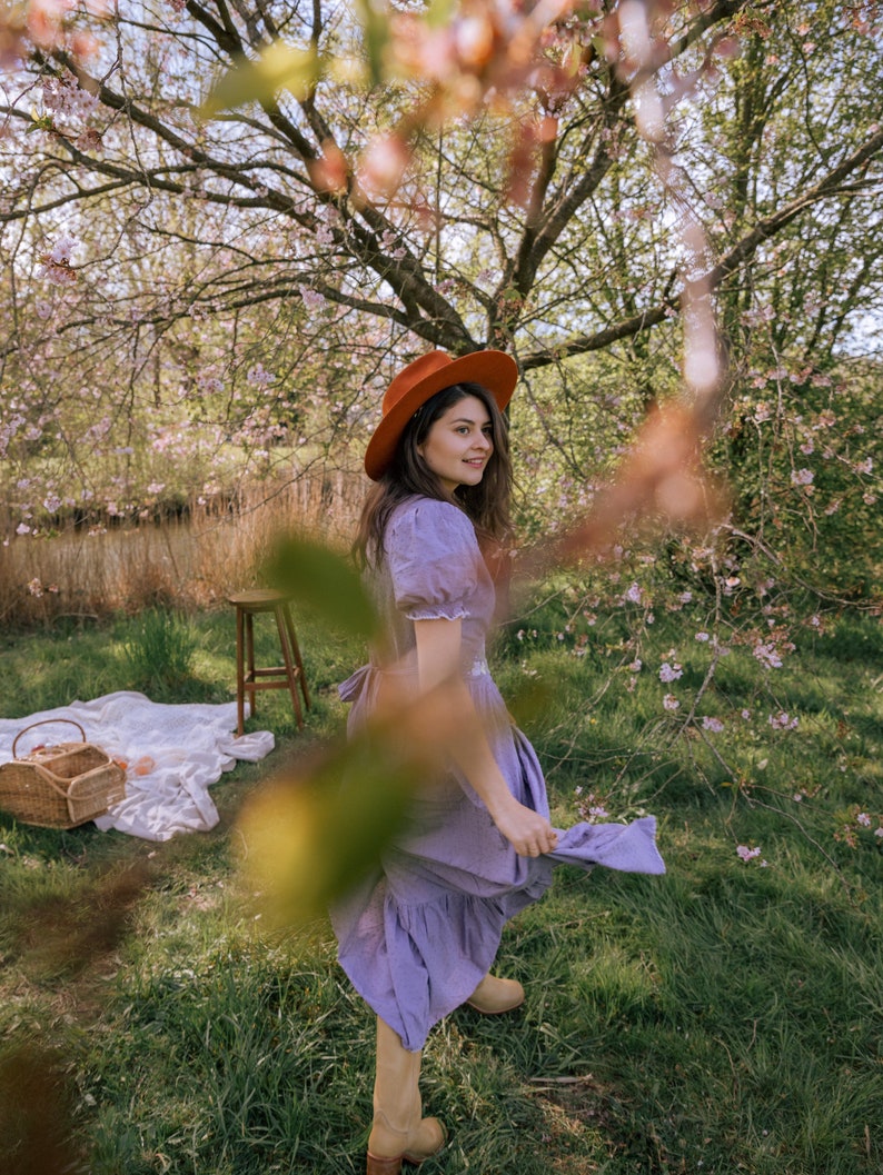 May include: A woman wearing a rust-colored hat and a lavender dress stands in a grassy area with a picnic basket and blanket. The scene is framed by a flowering tree, creating a soft, natural aesthetic. The dress has a ruffled hem and short sleeves.