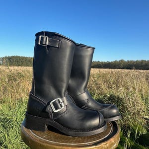 Könnte beinhalten: Ein Paar schwarze Motorradstiefel aus Leder mit silbernen Schnallen und Nähten. Die Stiefel sind kniehoch und haben eine runde Spitze und eine dicke Sohle. Sie werden auf einem kleinen, runden, messingfarbenen Tisch vor einem Feld und blauem Himmel präsentiert.