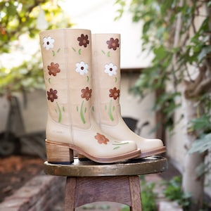 May include: A pair of beige leather boots with floral embroidery. The boots have a brown, white, and green floral pattern. The boots are sitting on a wooden stool.