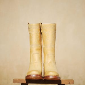 Puede incluir: Un par de botas de vaquero de color beige claro con costuras decorativas. Las botas son de cuero y tienen un dise&ntilde;o occidental cl&aacute;sico. Se exhiben sobre un peque&ntilde;o taburete de madera contra un fondo neutro.