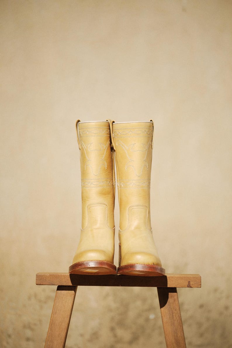 Puede incluir: Un par de botas de vaquero de color beige claro con costuras decorativas. Las botas son de cuero y tienen un dise&ntilde;o occidental cl&aacute;sico. Se exhiben sobre un peque&ntilde;o taburete de madera contra un fondo neutro.