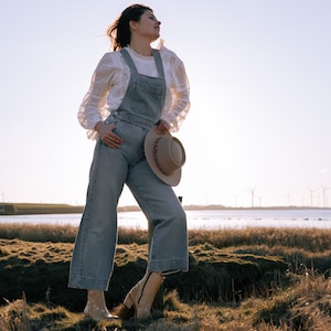 May include: A woman wearing a white lace blouse and blue denim overalls stands in a field of tall grass. She is holding a brown hat in her hand and looking up at the sky.