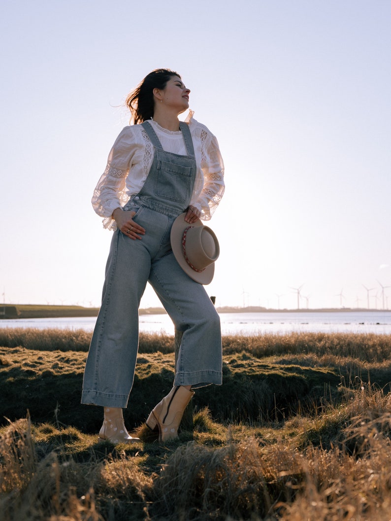 May include: A woman wearing a white lace blouse and blue denim overalls stands in a field of tall grass. She is holding a brown hat in her hand and looking up at the sky.