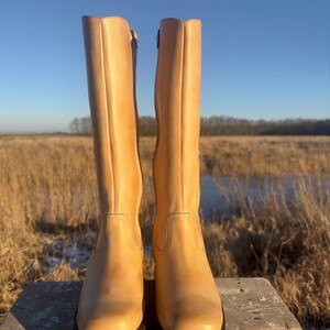 May include: A pair of tall, light brown leather boots with a chunky heel and a square toe. The boots are sitting on a wooden stool against a backdrop of a field and a blue sky.