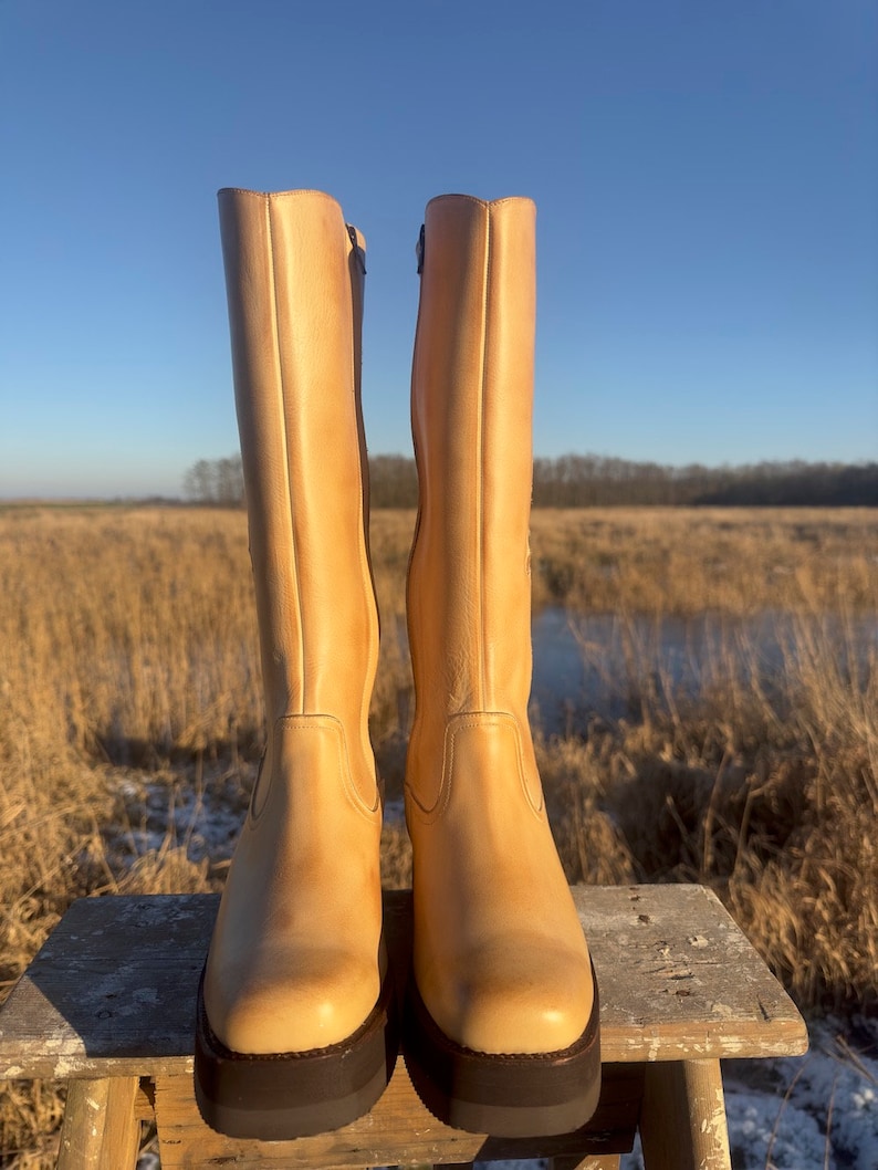 May include: A pair of tall, light brown leather boots with a chunky heel and a square toe. The boots are sitting on a wooden stool against a backdrop of a field and a blue sky.