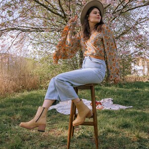 May include: A woman wearing a floral print blouse, blue jeans, and brown ankle boots sits on a wooden stool in a grassy field. She is wearing a wide-brimmed hat and looking off to the side.