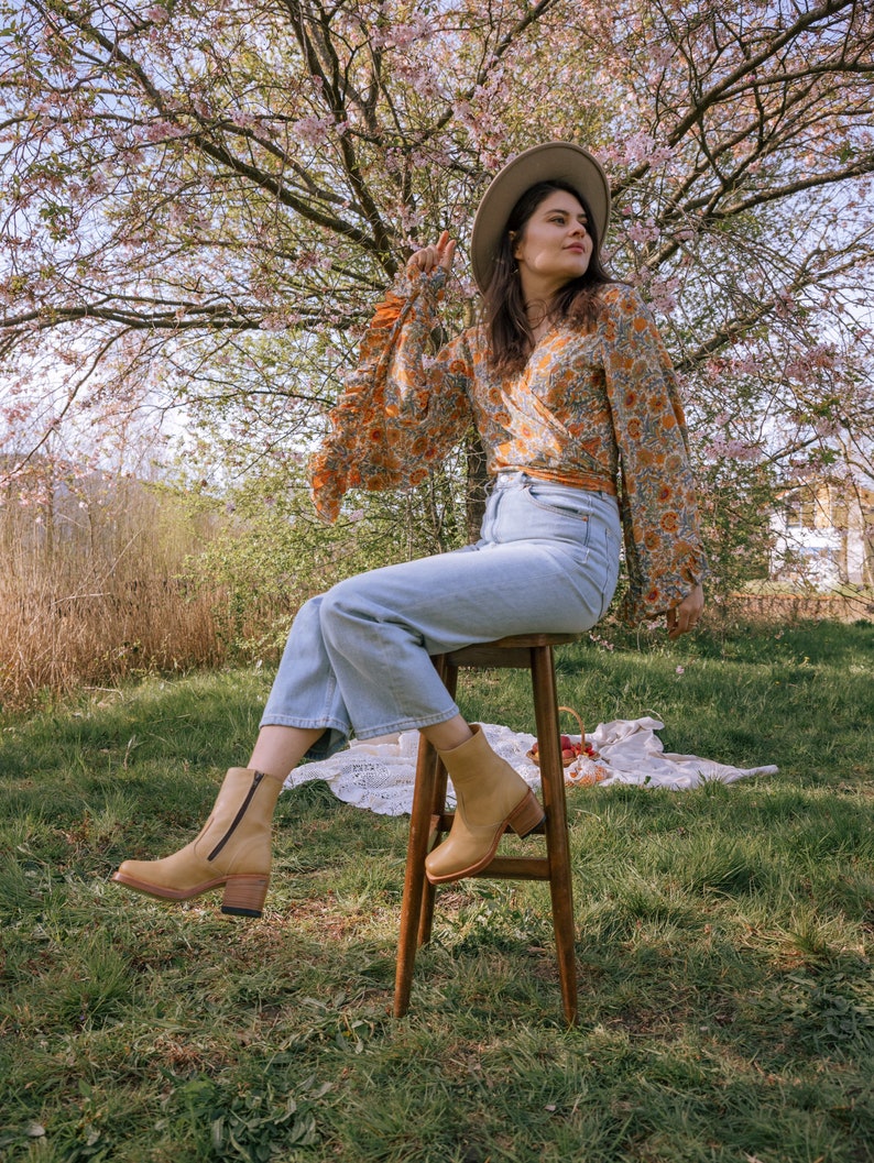 May include: A woman wearing a floral print blouse, blue jeans, and brown ankle boots sits on a wooden stool in a grassy field. She is wearing a wide-brimmed hat and looking off to the side.