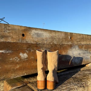 Puede incluir: Un par de botas de ante beige con tac&oacute;n de madera apilada. Las botas tienen ca&ntilde;a hasta la rodilla. Se colocan sobre tablas de madera desgastadas y un cielo azul claro.