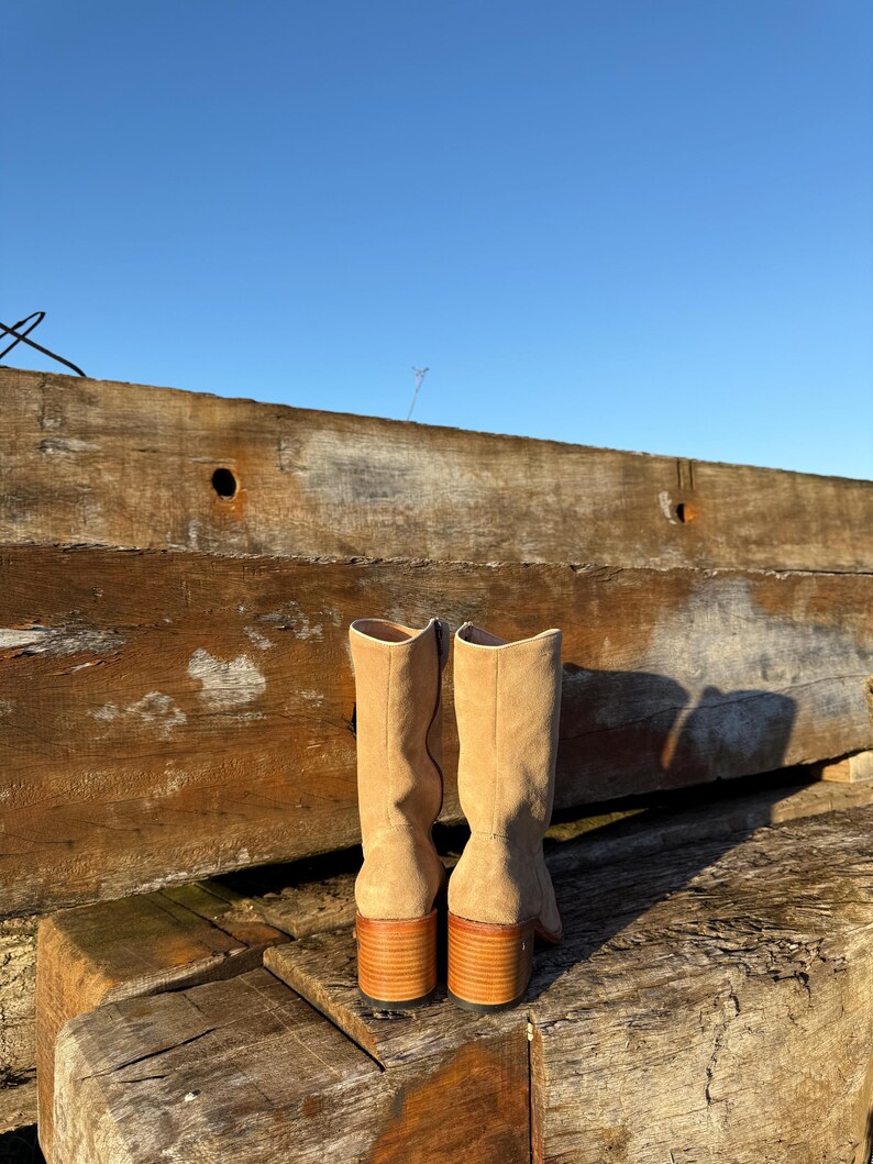 Puede incluir: Un par de botas de ante beige con tac&oacute;n de madera apilada. Las botas tienen ca&ntilde;a hasta la rodilla. Se colocan sobre tablas de madera desgastadas y un cielo azul claro.