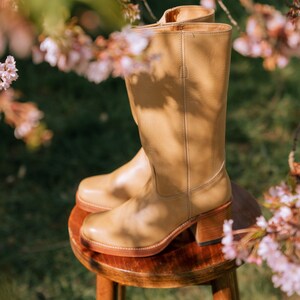 May include: A pair of tan leather knee-high boots with a block heel, resting on a small wooden stool. The boots have a classic design with a rounded toe and a zipper closure. The background features green grass and pink blossoms.