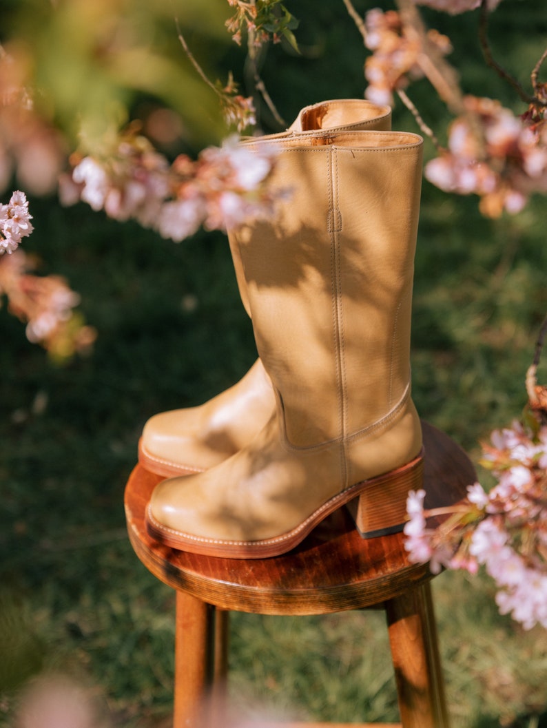 May include: A pair of tan leather knee-high boots with a block heel, resting on a small wooden stool. The boots have a classic design with a rounded toe and a zipper closure. The background features green grass and pink blossoms.