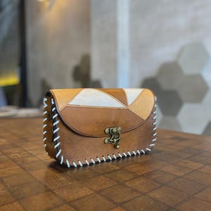 Op de afbeelding: Bruine leren crossbodytas met een geometrisch ontwerp op de flap. De tas heeft een messing sluiting en witte stiksels langs de randen. De tas staat op een houten oppervlak.