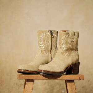 May include: A pair of beige suede ankle boots with decorative white stitching. The boots have a zipper closure, a rounded toe, and a stacked heel. They are displayed on a small wooden stool.