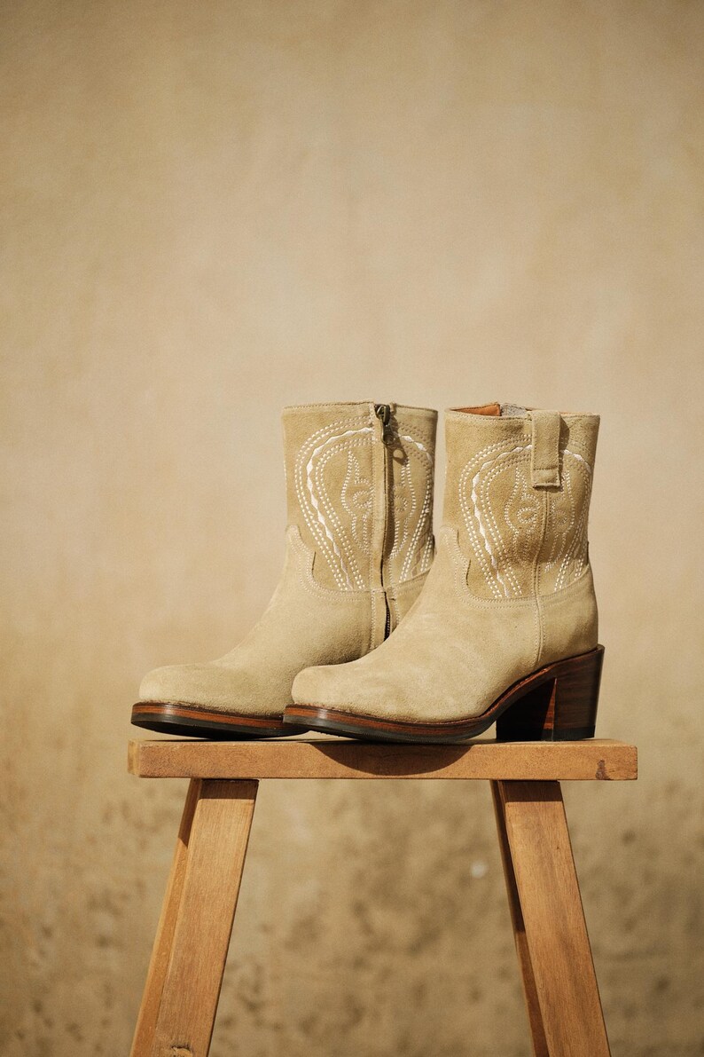 May include: A pair of beige suede ankle boots with decorative white stitching. The boots have a zipper closure, a rounded toe, and a stacked heel. They are displayed on a small wooden stool.