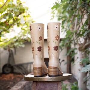 May include: A pair of beige leather boots with floral embroidery. The boots have a block heel and are sitting on a wooden stool.