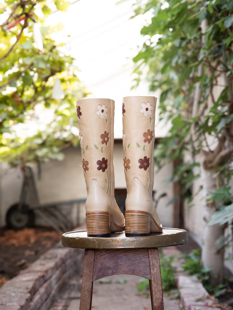 May include: A pair of beige leather boots with floral embroidery. The boots have a block heel and are sitting on a wooden stool.