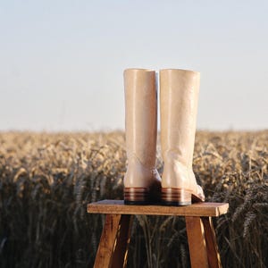 May include: A pair of beige leather boots with a low heel, standing on a small wooden stool. The boots are set against a backdrop of a wheat field under a clear sky, suggesting a rustic or countryside theme.